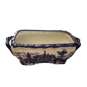 May include: Rectangular blue and white ceramic planter with a landscape design. The planter has a scalloped rim and handles on each end. The blue design depicts buildings and trees. The interior is a cream color.