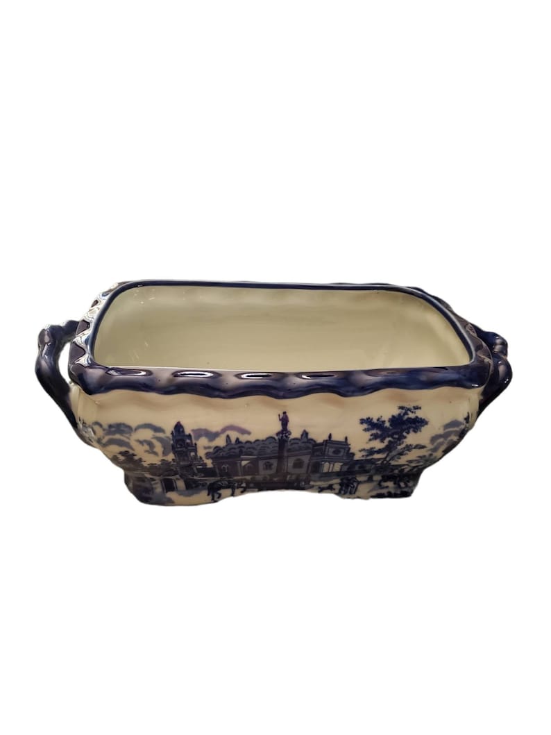 May include: Rectangular blue and white ceramic planter with a landscape design. The planter has a scalloped rim and handles on each end. The blue design depicts buildings and trees. The interior is a cream color.