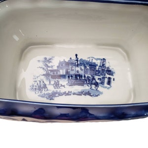 May include: A rectangular, ceramic serving dish with a blue and white transferware design. The dish features a scalloped rim and a detailed scene of a building and figures. The interior is white, and the rim is a deep blue.
