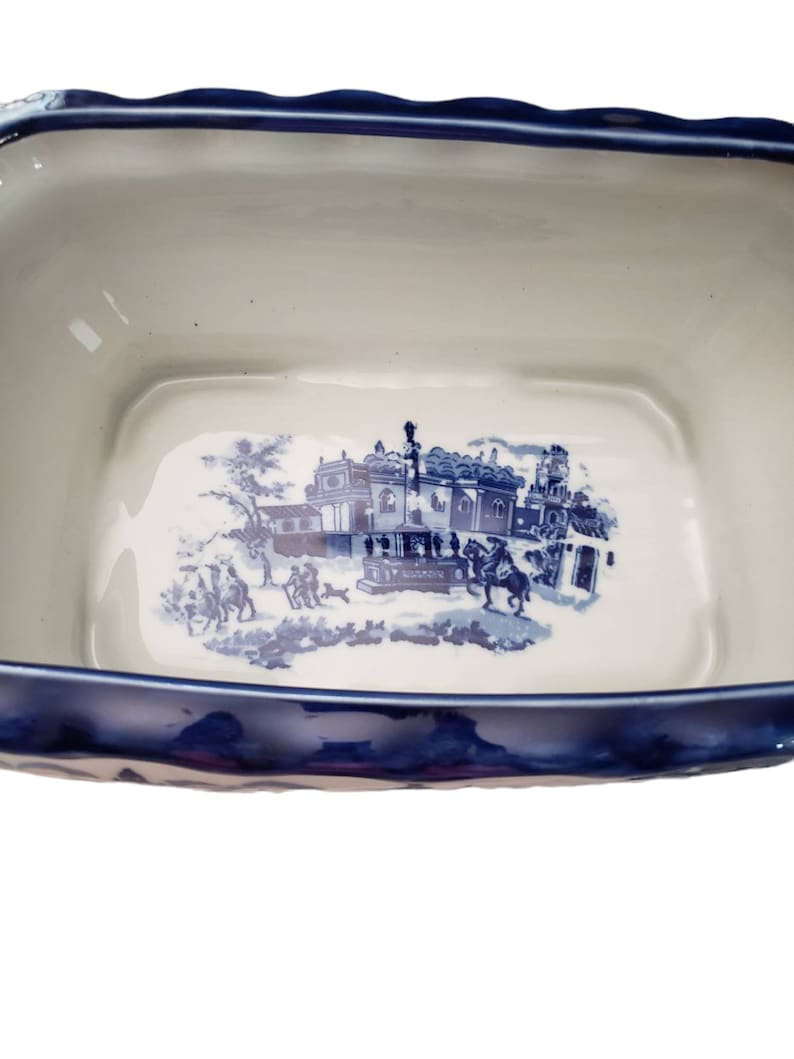 May include: A rectangular, ceramic serving dish with a blue and white transferware design. The dish features a scalloped rim and a detailed scene of a building and figures. The interior is white, and the rim is a deep blue.