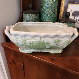 May include: White rectangular ceramic planter with a scalloped rim and handles. The planter features a green landscape design. Other green ceramic items are in the background, including a vase and two lidded jars. The planter sits on a wooden surface.