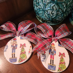 May include: Two round, white ceramic ornaments with a nutcracker and Christmas tree design. Each ornament is tied with a red and blue plaid ribbon with gold trim.