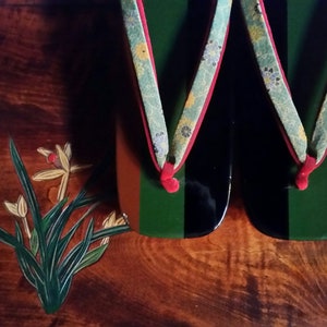 May include: A pair of traditional Japanese wooden sandals, known as geta, with black, green, and brown painted designs. The sandals have floral patterned straps in shades of blue and green.