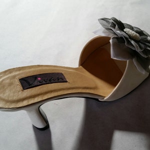 May include: White satin heeled slide with a black and white flower embellishment. The insole is tan leather with the brand name "Vivian" printed on it.
