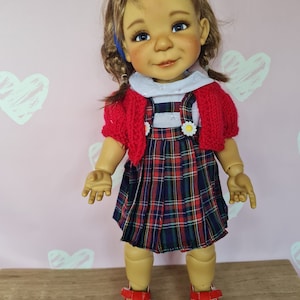 May include: A doll with a fair complexion, blue eyes, and freckles, dressed in a red cardigan, a white collared shirt, and a plaid dress. The doll has red shoes and a blue bow in her hair. The dress features a small daisy.