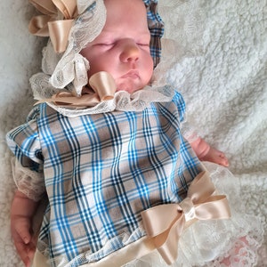 May include: A baby wearing a vintage-style outfit. The ensemble includes a plaid top in blue, tan, and white, paired with white lace-trimmed bloomers. A matching bonnet with lace and a large bow completes the look. The outfit is accented with satin bows.