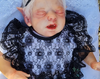 May include: A reborn doll with pale skin, closed eyes, and pointed ears. The doll wears a black and white lace dress with a black headband. The doll is lying on a white surface.