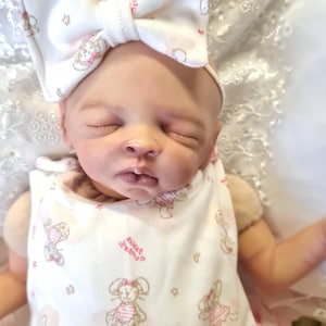May include: A realistic baby doll dressed in a white onesie and a matching bow headband, both featuring a pink and brown bunny design. The onesie is printed with the words "sweet dreams".