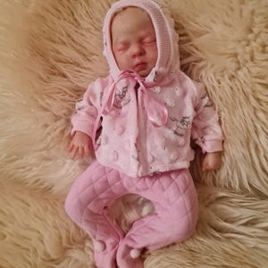 May include: A pink and white baby doll wearing a hooded jacket and quilted trousers. The doll is lying on a fluffy white surface.