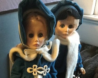 May include: Two dolls dressed in winter clothing stand on a white railing. The girl doll wears a blue coat with white trim and a purple skirt. The boy doll wears a blue coat with a white scarf. The dolls are standing on a white railing with a wooden base.