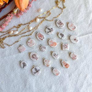 May include: A gold-toned chain necklace with small white pearls and a collection of small, white, oval and shaped charms featuring various illustrated woodland animals, including foxes, raccoons, owls, and birds in soft, muted watercolor tones.