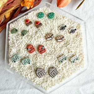 May include: A collection of novelty earrings in a clear hexagonal container filled with white rice. The earrings feature various designs, including cameras, backpacks, tents, cacti, houses, and campfires. The earrings are in various colors.