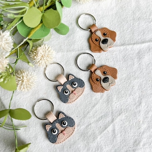 Puede incluir: Cuatro llaveros con caras de animales: dos gatos y dos perros. Los llaveros de gato tienen caras grises y beige, mientras que los llaveros de perro son marrones y blancos. Cada llavero tiene un anillo de metal plateado.