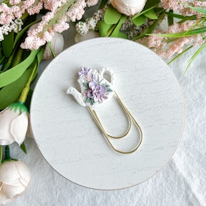 May include: White ceramic teapot bookmark with purple and green floral details. The teapot is attached to a gold metal bookmark clip.