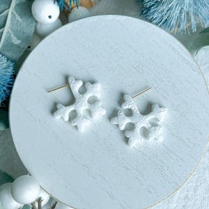 May include: Two white snowflake stud earrings with gold-coloured posts. The earrings are presented on a white, round surface, with blue and white decorative elements in the background. Ideal for winter or festive occasions.