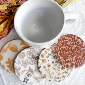 Könnte beinhalten: Ein weißer Keramikbecher über vier runden Untersetzern mit verschiedenen Mustern: eines mit gelben Eichenblättern und Eicheln, eines mit beigen Blumenmotiven, eines mit kleinen Wildblumen und eines mit rostfarbenem Farnblattmuster.