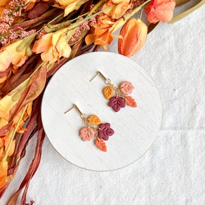 May include: A pair of gold-tone earrings featuring clusters of colorful leaf charms in shades of orange, red, and peach. The earrings are displayed on a white circular surface, with fall-themed floral accents in the background.