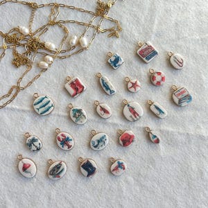 May include: A collection of nautical-themed charms with gold-colored loops, featuring various designs like fish, anchors, and lobsters. A gold chain with pearls is also present. The charms are white with colorful illustrations, perfect for jewelry making or crafting.