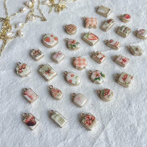 Op de afbeelding: Een verzameling kleine, decoratieve bedels met goudkleurige randen en diverse ontwerpen, waaronder aardbeien, bloempatronen en ruitpatronen. Er is ook een goudkleurige ketting met parels aanwezig.