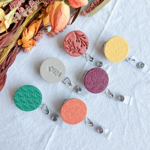 May include: A collection of six round badge reels in various colors, including green, orange, and yellow. Each reel features a unique embossed design, such as leaves or floral patterns. The reels are displayed on a white surface, with dried flowers in the background.