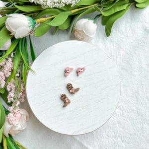 May include: A pair of pink flower stud earrings and two pairs of brown cowboy boot earrings with pink flower accents are displayed on a white circular surface. The background includes white and pink flowers and green foliage.