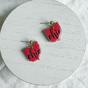 Può includere: Un paio di orecchini rossi a forma di Ohio con ganci dorati e la parola "Ohio" scritta in corsivo nero.