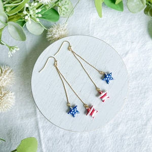 May include: A pair of gold-tone dangle earrings. Each earring features a delicate chain suspending a star charm. One star is blue with white polka dots, the other is red with white stripes. The earrings are displayed on a white surface.