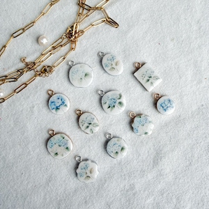 May include: A collection of white ceramic pendants with blue floral designs, scattered on a light-colored surface. A gold chain and several pearls are also present. The pendants vary in shape, including round, oval, and square.