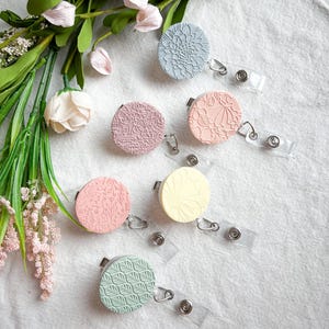 May include: Six pastel-coloured badge reels with embossed floral and geometric patterns. The reels are in shades of blue, pink, yellow, and green, with clear plastic clips and retractable cords. Greenery and flowers are arranged in the background.