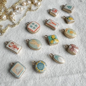 May include: A collection of small, decorative pendants with various designs, including a sailboat, lobster, and lemons. The pendants are gold-toned with colorful enamel details. A gold chain with pearls is also present.
