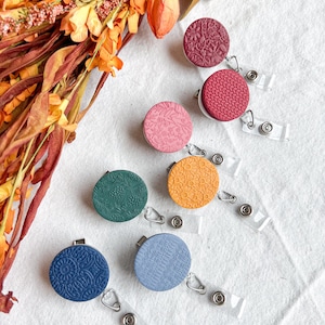 Puede incluir: Una colección de portatarjetas de colores y con diseños. Los carretes son redondos y presentan varios diseños en relieve en tonos de rosa, rojo, verde, naranja, azul y azul claro. Cada carrete tiene un cordón retráctil de plástico transparente y un clip metálico.