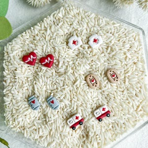 May include: A collection of miniature medical-themed stud earrings. Designs include red hearts with white lines, nurse hats with red crosses, bandages with hearts, ambulance cars with red crosses, and medical bags with hearts. The earrings are displayed on a bed of white rice.