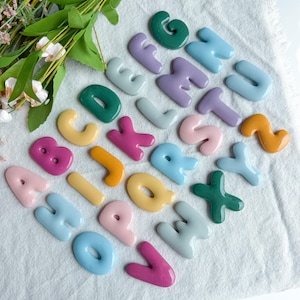May include: A collection of twenty-six individual alphabet letters made of smooth, glossy material in various colors including pink, yellow, green, blue, and purple. Each letter is shaped with rounded edges and a shiny finish, arranged in a scattered layout.