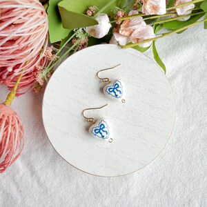 May include: A pair of heart-shaped earrings with a white base and blue bow design. The earrings have gold-colored hooks and are displayed on a white, round surface. Floral accents in pink and green are in the background.