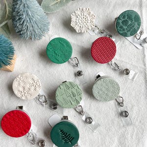 May include: Nine different Christmas themed retractable badge holders. The holders are round and feature various designs including snowflakes, pine trees, and a knit pattern. The holders are attached to clear plastic retractable cords.