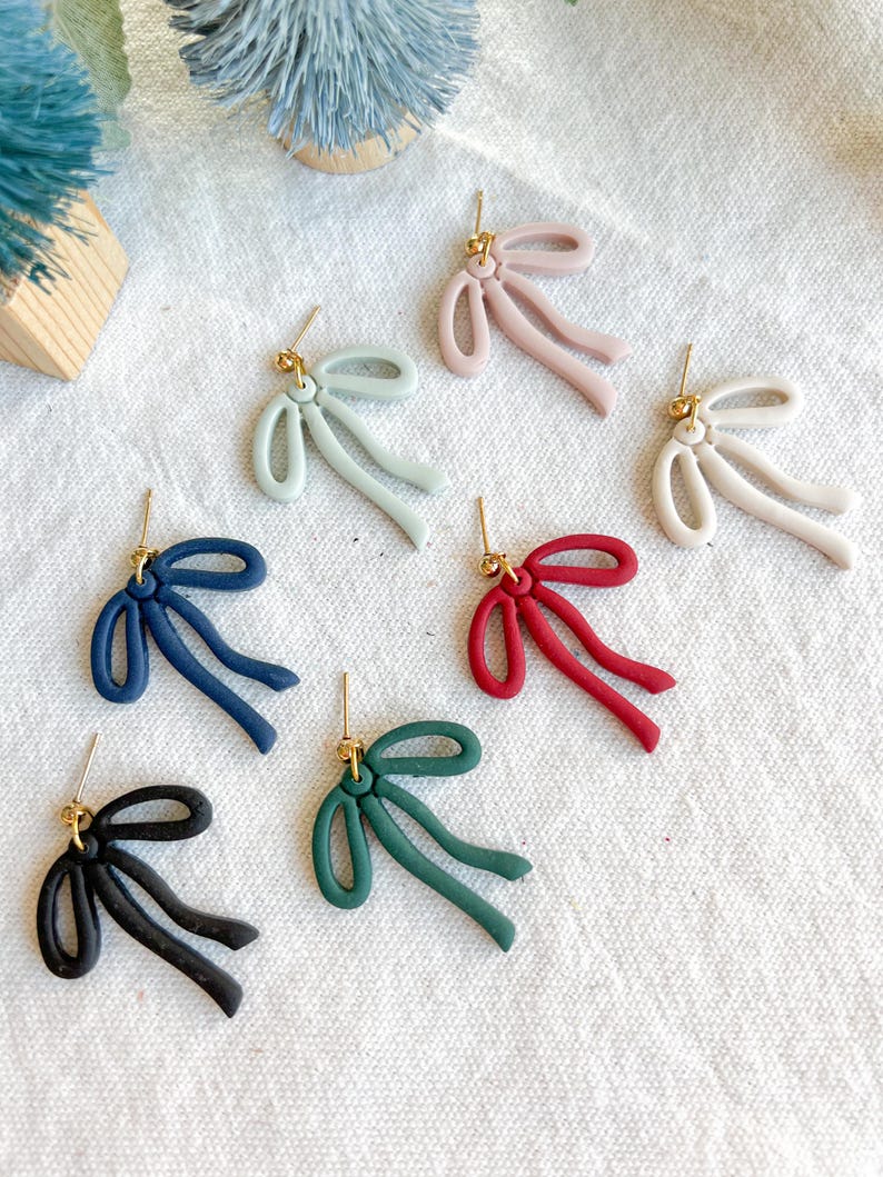 May include: Assortment of six pairs of bow-shaped earrings in various colors, including black, navy blue, green, red, light blue, and pink. The earrings are made of polymer clay and have a gold-tone post.