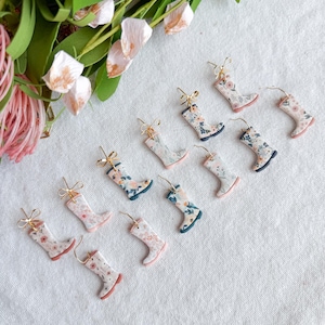 Puede incluir: Un conjunto de pendientes con forma de botas de agua con estampado floral, con ganchos dorados. Las botas presentan varios diseños florales en rosa, azul y blanco, con lazos dorados en la parte superior. Dispuestas sobre una superficie blanca.