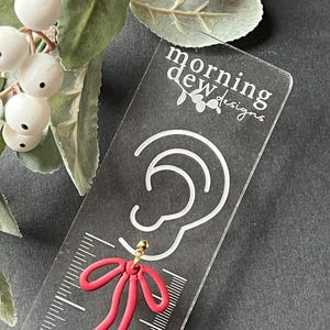 May include: Red bow earring with a gold post, displayed on a clear acrylic card with the text "morning dew designs".