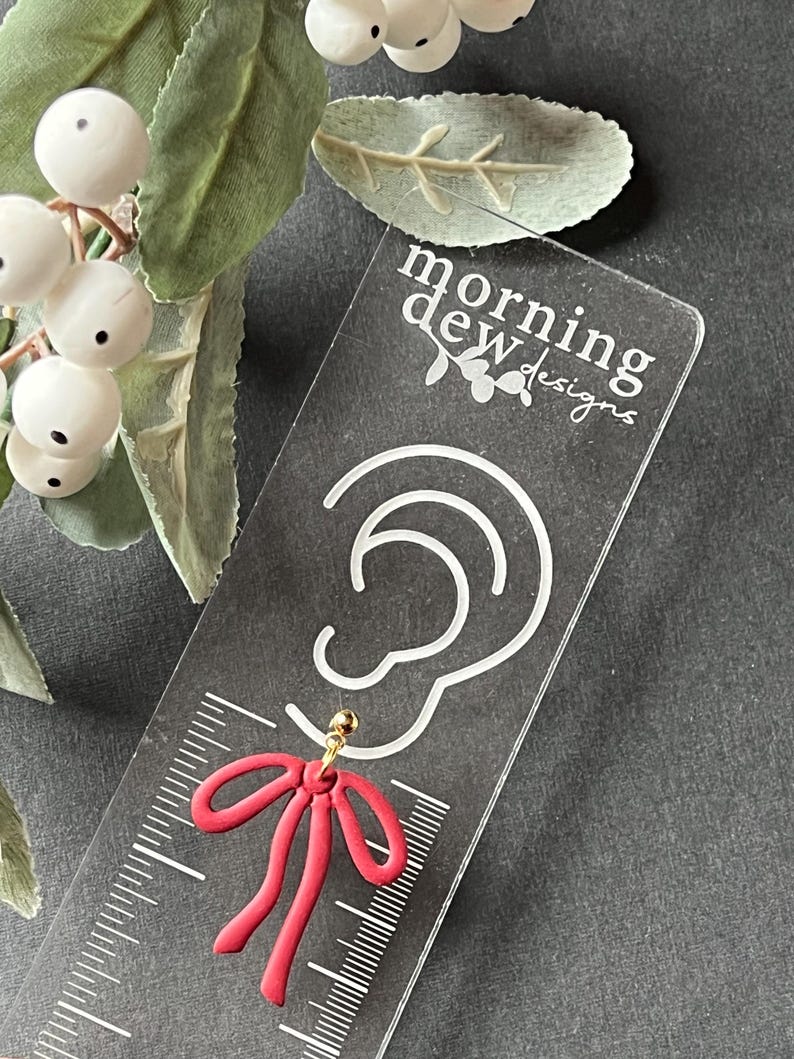 May include: Red bow earring with a gold post, displayed on a clear acrylic card with the text "morning dew designs".
