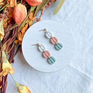 May include: A pair of dangle earrings with three clay beads in shades of white, pink, and green. The beads are shaped like rounded rectangles and have a textured surface.