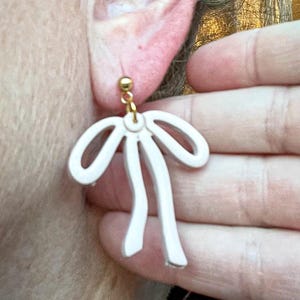 May include: A pair of white bow-shaped earrings with gold accents. The earrings are hanging from the earlobe of a person.