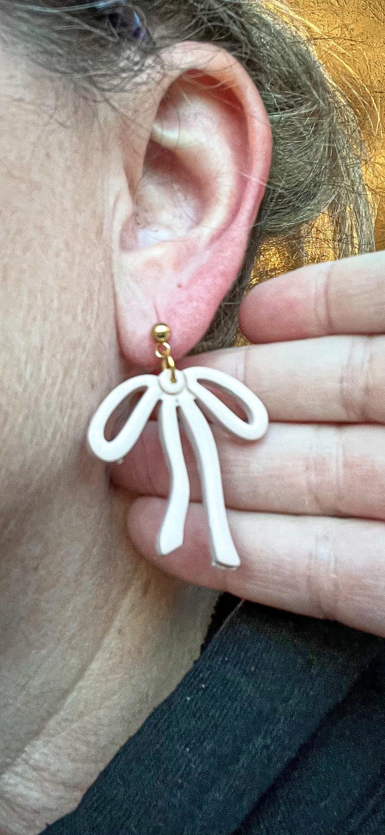 May include: A pair of white bow-shaped earrings with gold accents. The earrings are hanging from the earlobe of a person.