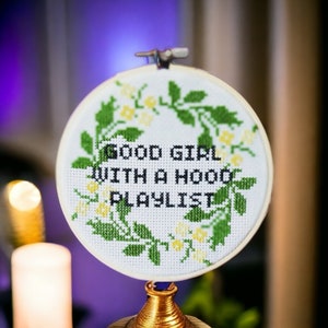 May include: A white embroidery hoop with a green floral wreath and the text "Good Girl With A Hood Playlist".