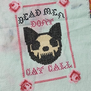 May include: Cross-stitch design on pale blue fabric featuring a black and cream cat skull. The text reads "DEAD MEN DON'T CAT CALL" in black and pink thread. Pink roses are in each corner.