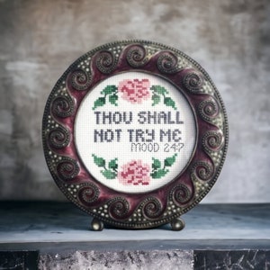 Op de afbeelding: Een decoratief rond bord met een bordeauxrode rand en een wit geborduurd ontwerp met een bloemenkrans en de tekst "Thou Shall Not Try Me Mood 24/7".