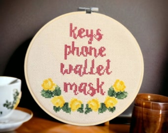 May include: A white embroidery hoop with a beige fabric background. The fabric has red embroidery that reads "keys phone wallet mask" and a border of yellow flowers with green leaves.