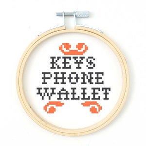 May include: A white embroidery hoop with the text "KEYS PHONE WALLET" in black pixelated font. Two orange pixelated phone icons are above and below the text.