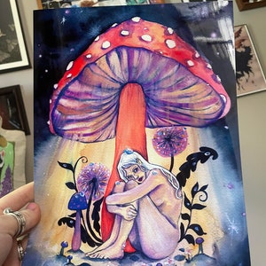 May include: A watercolor painting of a nude woman sitting under a large, red and purple mushroom with white spots. The woman has long, white hair and is holding her knees. The background is a dark blue with stars and a few small, purple mushrooms.