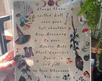 May include: A framed print with watercolor illustrations and text. The text reads "Always throw spilled salt over your left shoulder..." with images of a witch's hat, boots, a broom, and flowers. The print is held by a person.