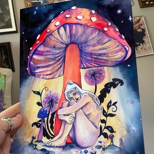 May include: A watercolor painting of a woman sitting under a large, red and purple mushroom with white spots. The woman is nude and has white hair and blue eyes. The background is a dark blue with white stars.